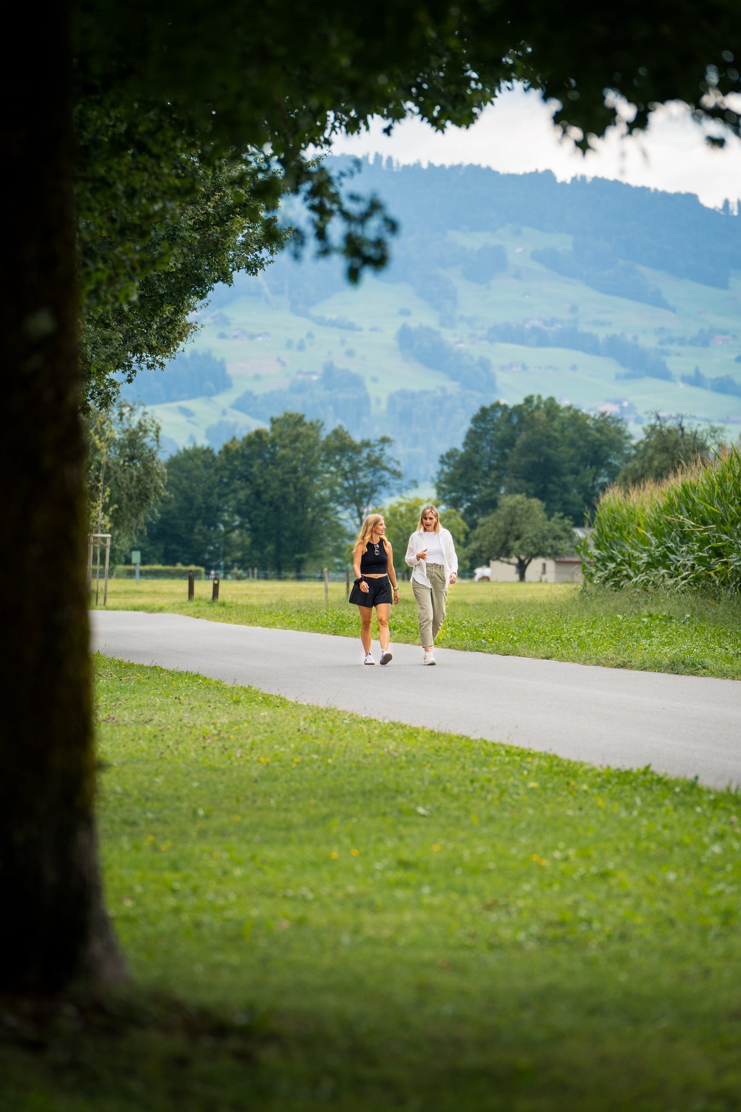 Zwei Frauen spazieren auf einem Weg durch eine grüne Landschaft, Goldenbody Shop Ch.