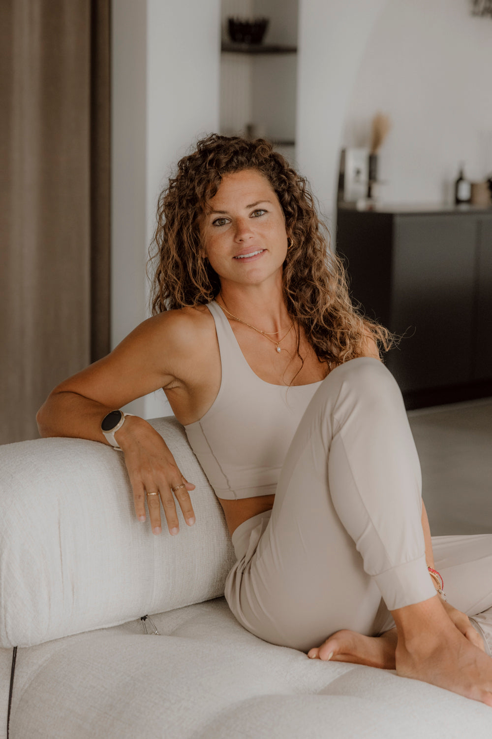 Eine Frau in beiger Sportkleidung sitzt entspannt auf einem Sofa von Goldenbody Shop Ch.
