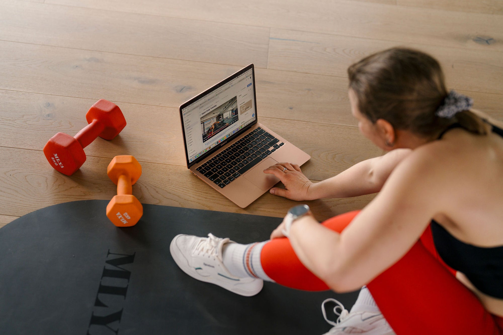 Frau trainiert mit Hanteln und Laptop von Goldenbody Shop Ch auf Fitnessmatte.