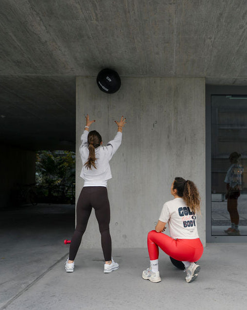 Zwei Frauen trainieren mit Medizinball, Goldenbody Shop Ch, unter Betonüberdachung.